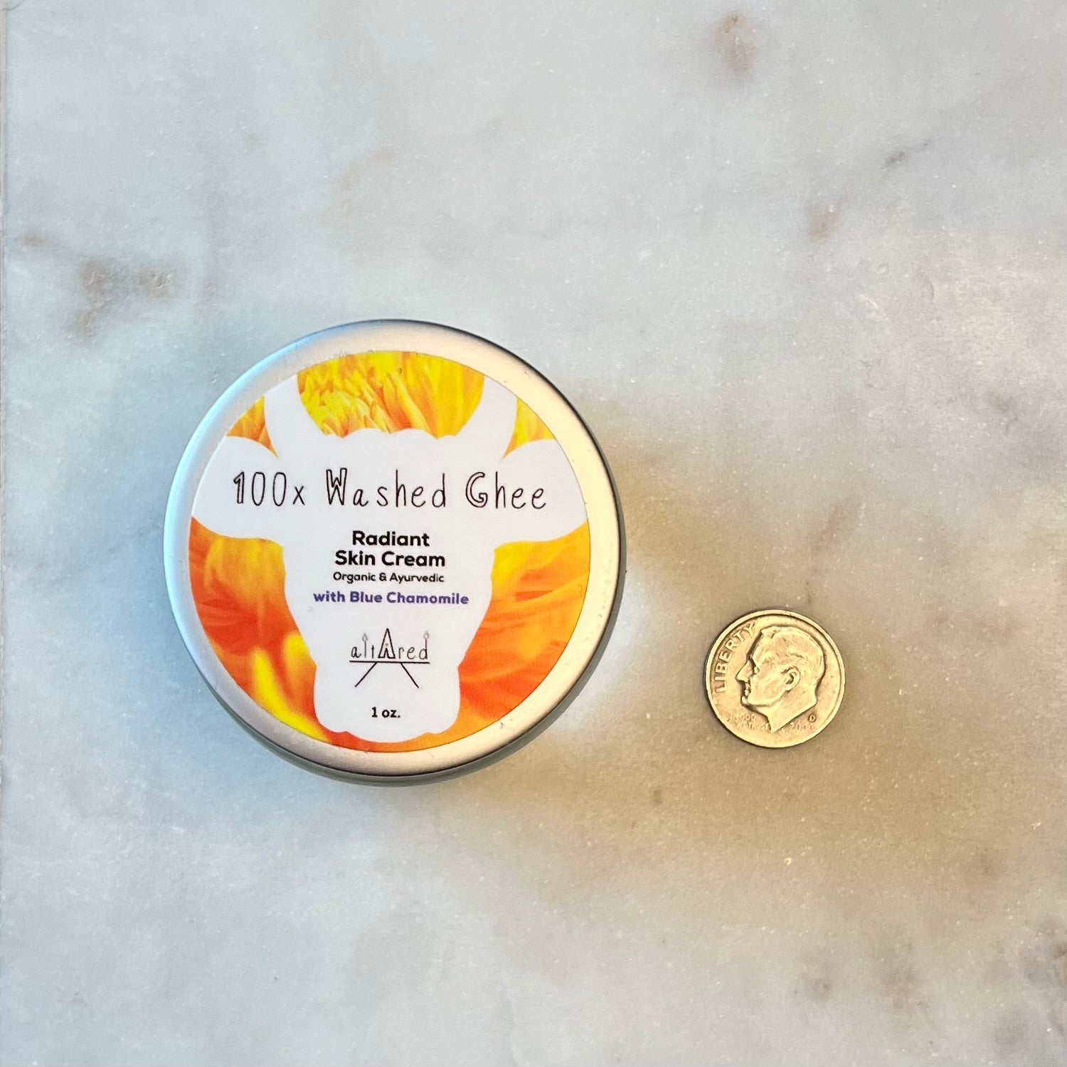 Top down 1 oz glass jar of 100x Washed Ghee Ayurvedic Skin Cream with German Chamomile on a marble board with a dime next to it for size comparison.
