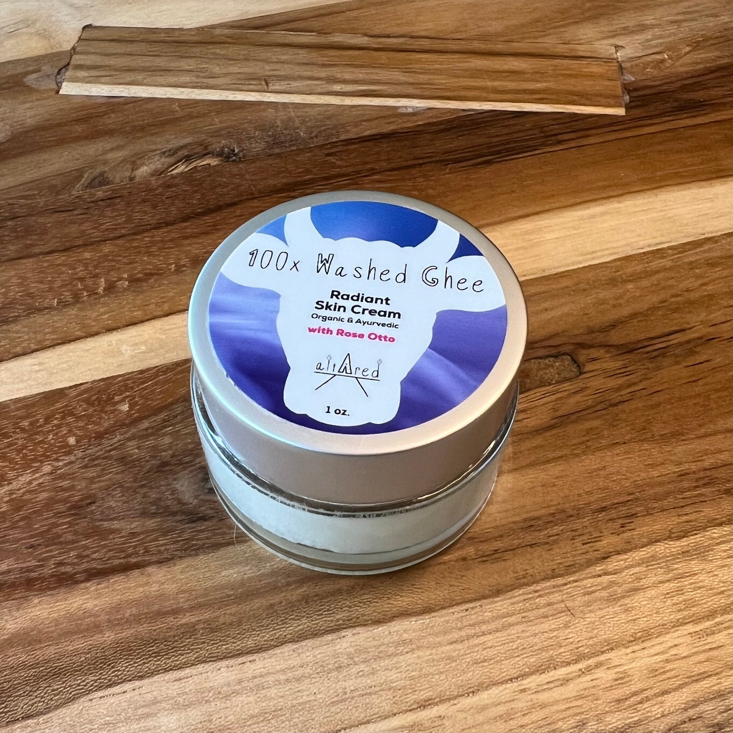 Jar of 1oz. 100x Washed Ghee Ayurvedic Skin Cream (Shata Dhauta Ghrita) with Rose Otto essential oil in a clear glass jar with a silver lid, blue label on top. It sits on a wooden table.