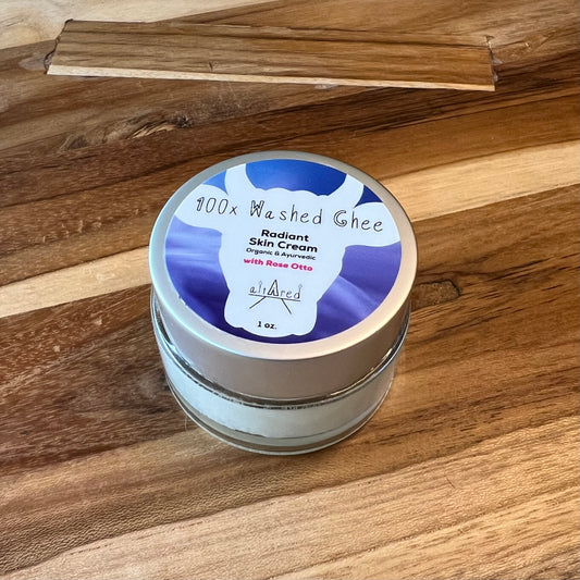 Jar of 1oz. 100x Washed Ghee Ayurvedic Skin Cream (Shata Dhauta Ghrita) with Rose Otto essential oil in a clear glass jar with a silver lid, blue label on top. It sits on a wooden table.