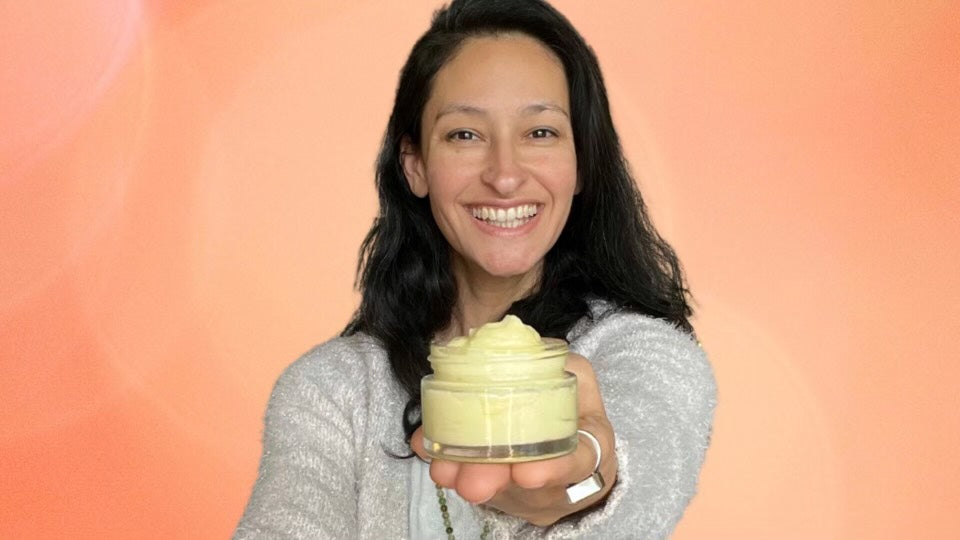 Amarylis holding out her jar of 100x Washed Ghee Ayurvedic Skin Cream toward the camera with a big smile against and orange background.