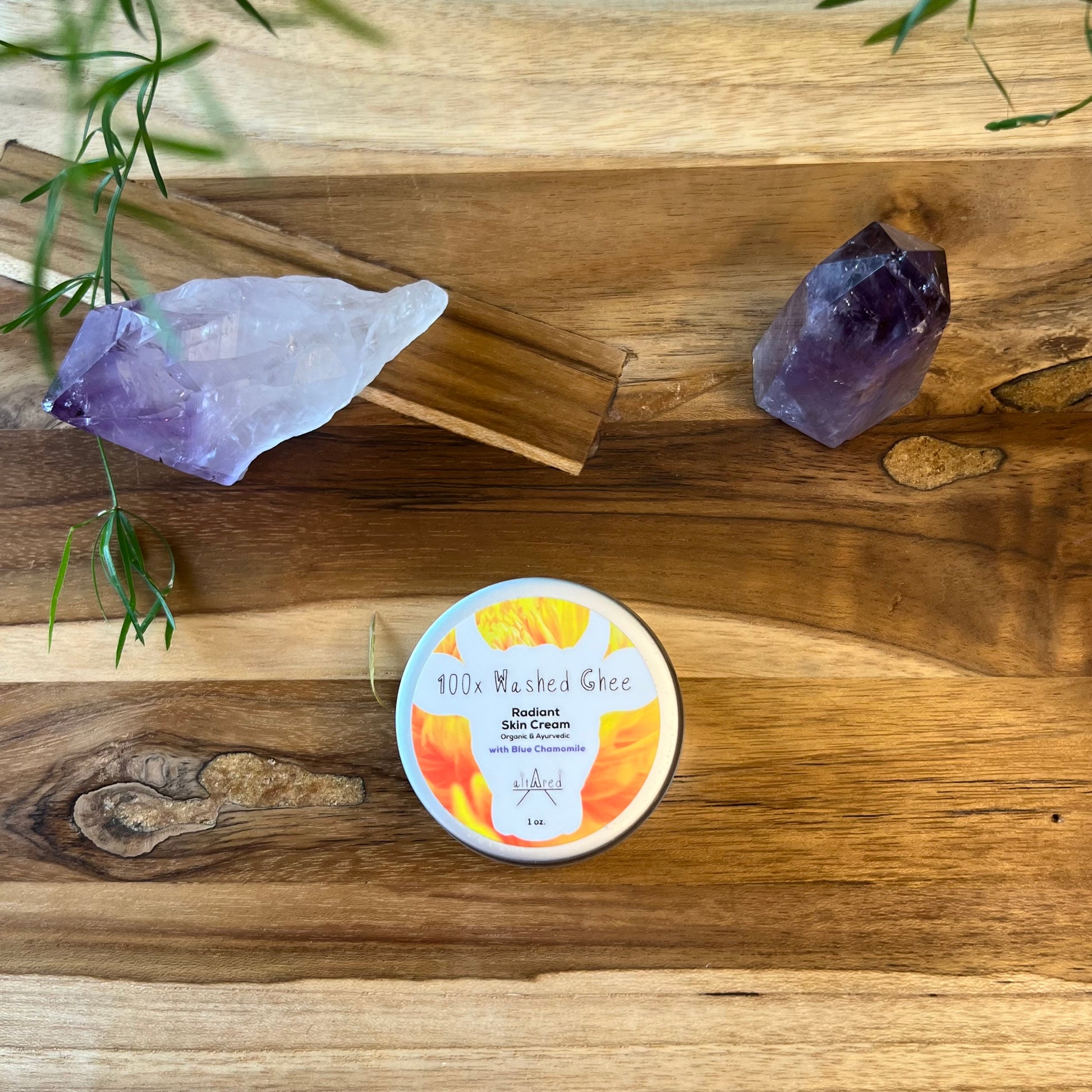 Top-down view of 1 oz. jar  of 100x Washed Ghee Ayurvedic Skin Cream with German Chamomile on a wooden board. Surrounded by two amethyst crystals.