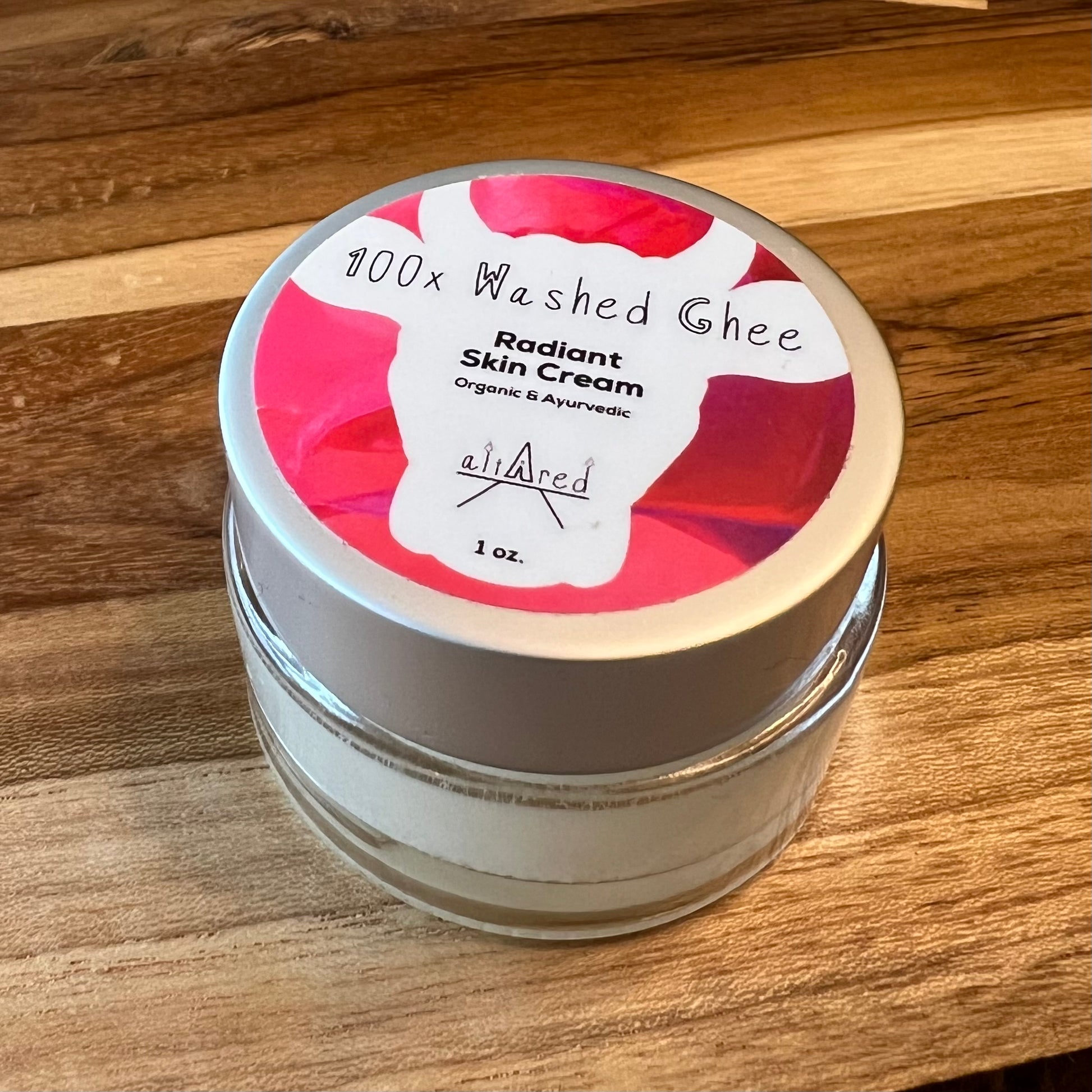 Jar of 100x Washed Ghee Ayurvedic Skin Cream (Shata Dhauta Ghrita) in a clear glass jar with a silver lid, pink label on top. It sits on a wooden table.